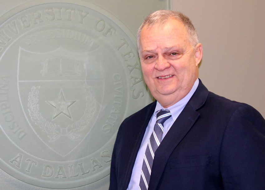 Office Bishop poses in a blue suit in front of a stone UT Dallas seal