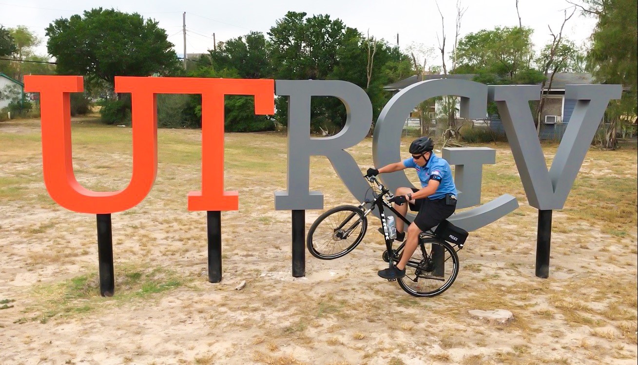 Office Ramos 'wheelies' on his bike in front of a sign composted of large letters 'UTRGV'